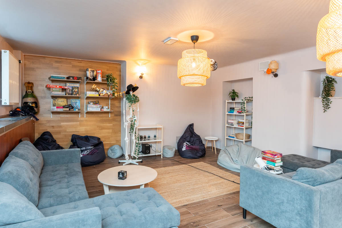 Przestrzeń wypoczynkowa z szarymi kanapami, szarymi i granatowymi pufami, regałami i półkami, na których ustawione są gry, książki.