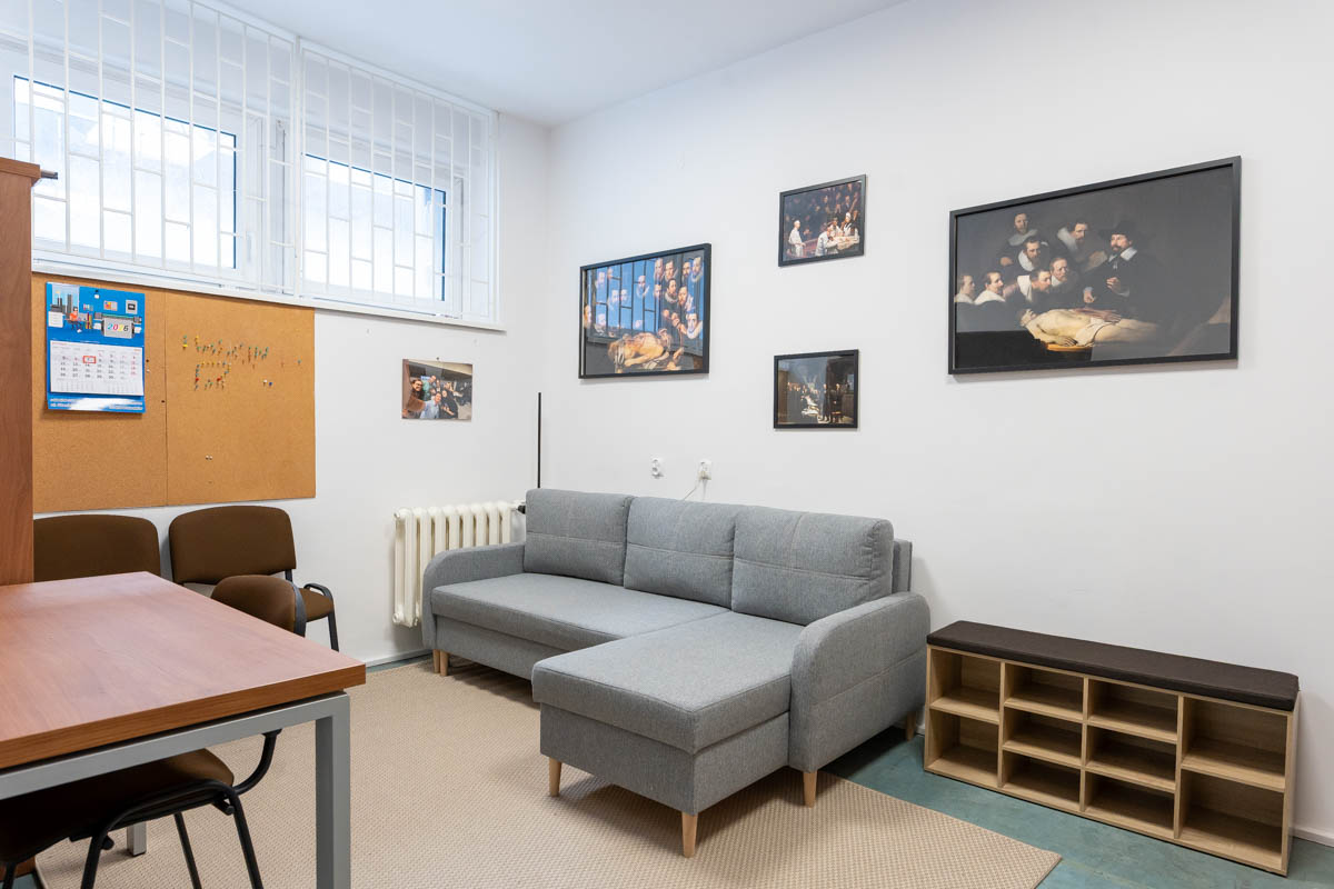 Pokój z szarą kanapą, regałem stojącym na ziemi, stołem i krzesłami. Na ścianie wiszą obrazy.