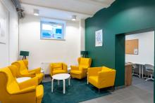 Przestrzeń wypoczynkowa z kilkoma żółtymi fotelami i małym fotelem ustawionymi wokół dwóch okrągłych stolików. Obok stoją dwie lampy. Na jednej ze ścian wisi obraz. W tle widać przejście do jasnego pomieszczenia z biurkami i krzesłami.