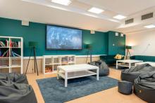 Strefa wypoczynkowa z kilkoma pufami i niskimi stolikami ustawionymi na dywanach. Na ścianie wisi duży ekran. Obok stoją regały z książkami i grami oraz lampy podłogowe. W tle znajdują się dwie kanapy z poduszką dekoracyjną. W pomieszczeniu nie ma osób.