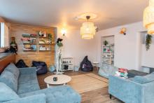 Przytulna sala z dwoma sofami, kilkoma pufami i niskimi stolikami ustawionymi na dywanie. Na ścianach stoją regały z książkami i grami. W rogu stoi model ludzkiego szkieletu. Delikatne oświetlenie i rośliny tworzą spokojną atmosferę. W pomieszczeniu nie ma osób.