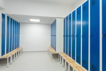Szatnia z dwoma rzędami wysokich szafek ustawionych w kształcie litery „L”. Przed szafkami stoją długie ławki. Pomieszczenie jest puste i dobrze oświetlone.