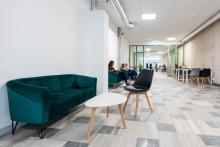 Korytarz z kanapą, stolikiem i krzesłem na pierwszym planie. W głębi osiem osób siedzi lub przechodzi dalej korytarzem. Część z nich korzysta z telefonów lub pracuje przy stolikach. Przestrzeń ma jasne oświetlenie i otwarty układ.