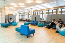 Przestrzeń wypoczynkowa z kilkoma osobami siedzącymi na pufach i niskich kanapach. Część osób korzysta z telefonów, a część rozmawia. W tle znajduje się strefa z biurkami i komputerami, przy których pracuje kilka osób. Pomieszczenie jest jasne i ma duże okna oraz ruchome ścianki, które dzielą przestrzeń.