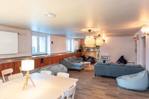 Przestrzeń wypoczynkowa z szarymi kanapami i szarymi i granatowymi pufami, małymi stolikami kawowymi, lampami zwisającymi z sufitu, białym stołem, przy którym stoją białe krzesła.