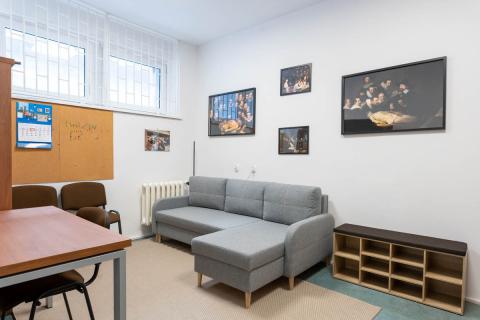 Pokój z szarą kanapą, regałem stojącym na ziemi, stołem i krzesłami. Na ścianie wiszą obrazy.