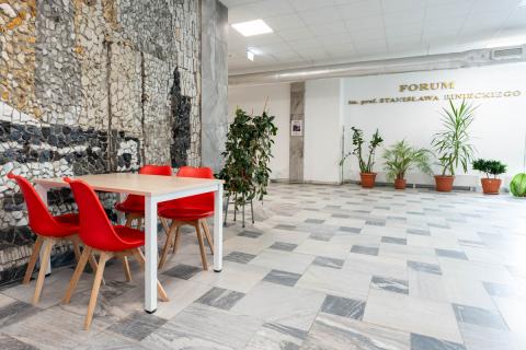 Przestrzeń Wydziału Farmaceutycznego, ze ścianą z mozaiki, zielonymi kwiatami doniczkowymi, czerwonymi krzesłami i stolikiem.